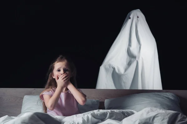 Niño asustado sentado en la cama y mostrando un signo de silencio mientras el fantasma blanco de pie detrás de la cama aislado en negro - foto de stock