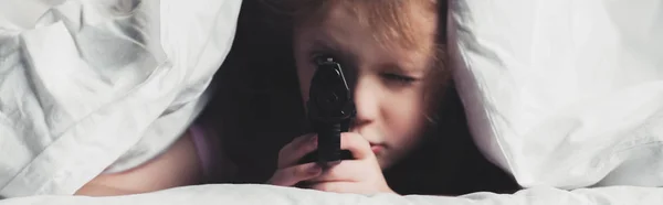 Panoramic shot of scared child holding gun while hiding under blanket — Stock Photo