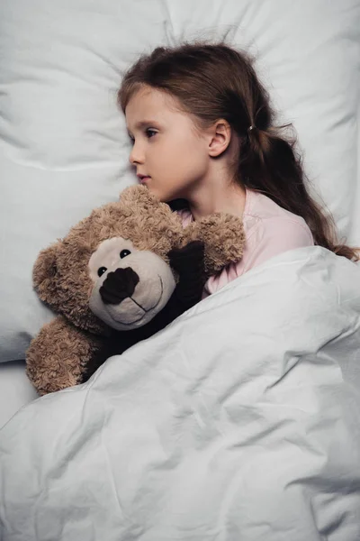 Adorável criança assustada deitada na cama com os olhos abertos perto de ursinho de pelúcia — Fotografia de Stock