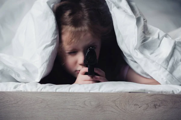 Verängstigtes Kind hält Pistole in der Hand, während es sich unter Decke im dunklen Schlafzimmer versteckt — Stockfoto