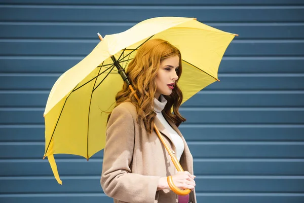 Приваблива жінка в пальто дивиться в сторону і тримає жовту парасольку — стокове фото