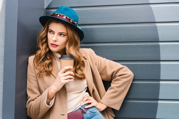 Attraente donna in cappello blu guardando lontano e tenendo tazza di carta — Foto stock