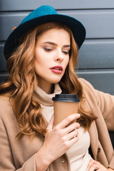 Attraente donna in cappello blu guardando lontano e tenendo tazza di carta — Foto stock