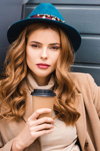 Atractiva mujer en sombrero azul mirando a la cámara y sosteniendo la taza de papel - foto de stock