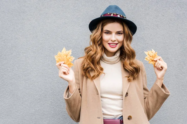 Attractive woman in blue hat looking at camera and holding yellow leaves — Stock Photo