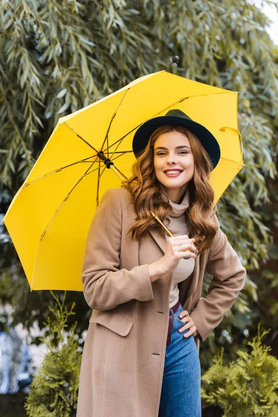 Attraente donna in cappello blu guardando la fotocamera e tenendo ombrello giallo — Foto stock