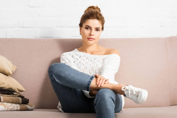 Attractive woman in white sweater looking at camera and sitting on sofa — Stock Photo