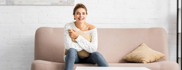 Panoramic shot of attractive woman in white sweater holding pillow and looking at camera — Stock Photo
