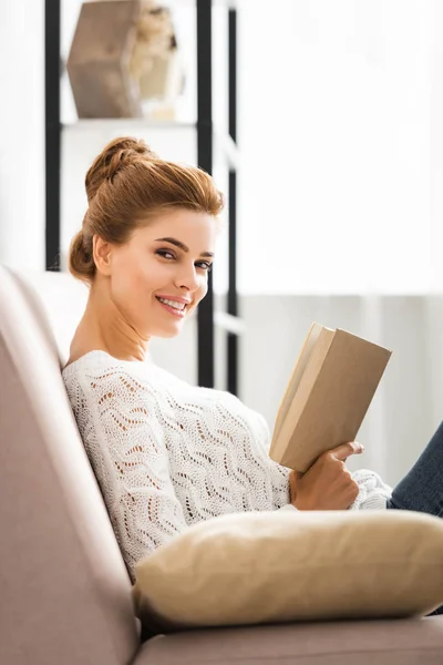 Attraktive Frau im weißen Pullover, Buch in der Hand und in die Kamera blickend — Stockfoto