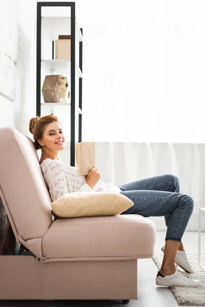 Attractive woman in white sweater holding book and looking at camera — Stock Photo