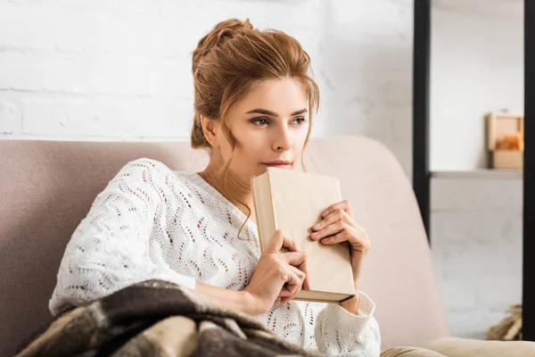Atractiva mujer en suéter blanco sosteniendo libro y mirando hacia otro lado - foto de stock