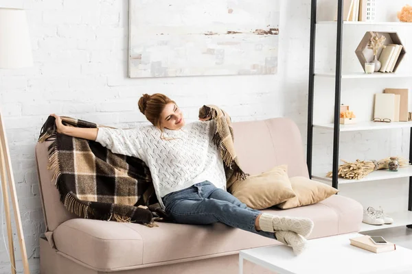 Femme attrayante avec couverture assise sur le canapé et regardant loin — Photo de stock