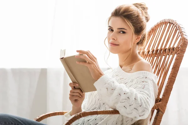 Привлекательная женщина в белом свитере сидит на кресле-качалке и держит книгу — стоковое фото