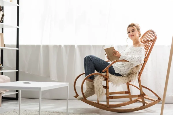 Mulher atraente em suéter branco sentado na cadeira de balanço e livro segurando — Fotografia de Stock