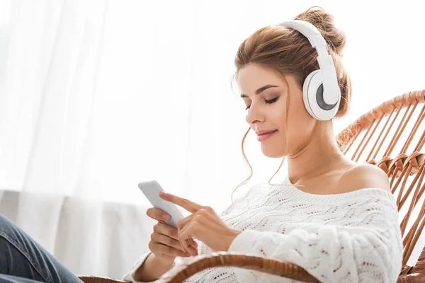 Atractiva mujer en suéter blanco escuchando música y usando un teléfono inteligente - foto de stock