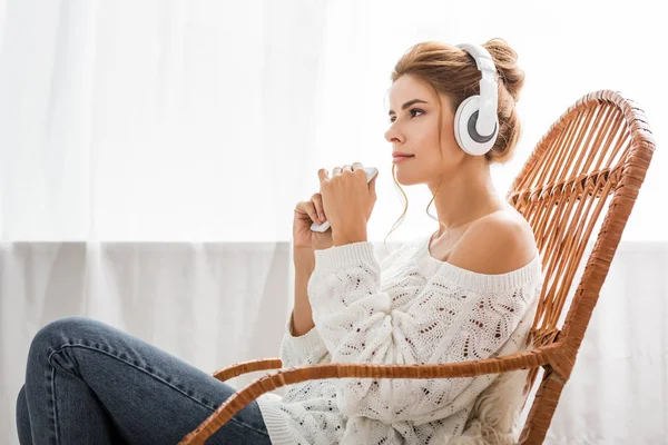 Atractiva mujer en suéter blanco escuchando música y sosteniendo el teléfono inteligente - foto de stock