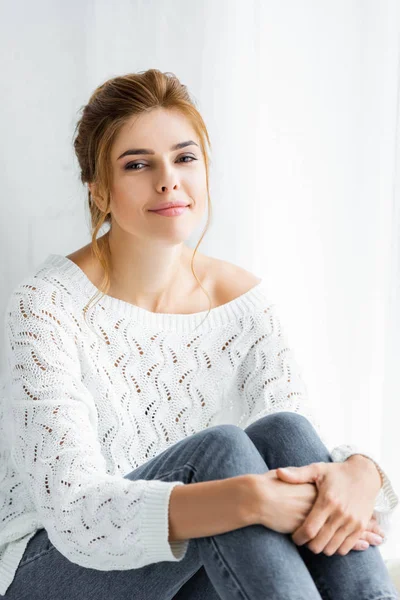 Jolie femme en pull blanc et jeans assis et regardant la caméra — Photo de stock