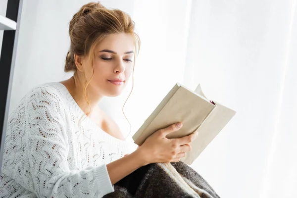 Atractiva mujer en suéter blanco con libro de lectura de manta - foto de stock