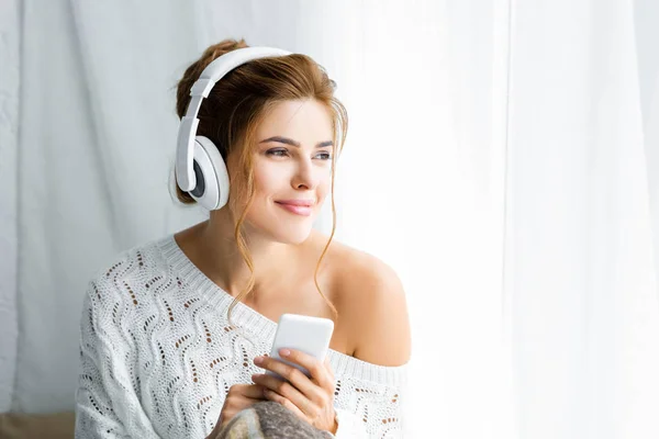 Приваблива жінка в білому светрі слухає музику і тримає смартфон — стокове фото