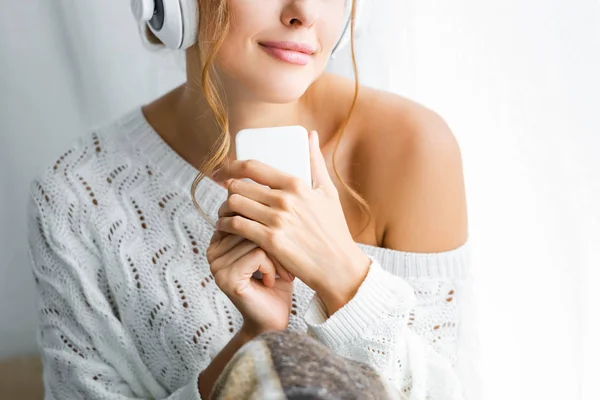 Vista recortada de la mujer en suéter blanco escuchando música y sosteniendo teléfono inteligente - foto de stock