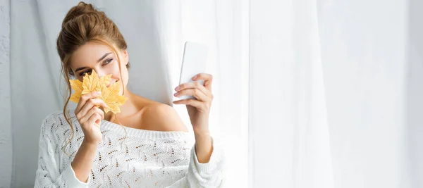 Prise de vue panoramique d'une jolie femme en pull blanc prenant des photos et tenant des feuilles jaunes — Photo de stock