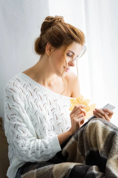 Attraktive Frau im weißen Pullover mit Smartphone und gelben Blättern — Stockfoto