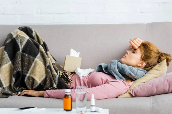 Вид збоку приваблива і хвора жінка в сірому шарфі з головним болем, що лежить в квартирі — стокове фото