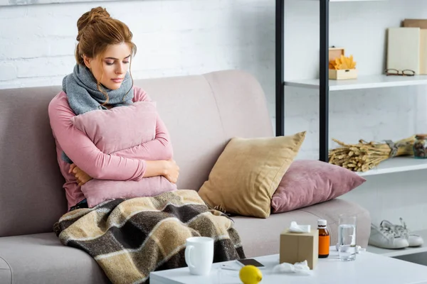 Mujer atractiva y enferma con bufanda gris sentada en el sofá y sosteniendo la almohada - foto de stock