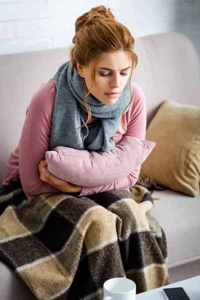 Mujer atractiva y enferma con bufanda gris sentada en el sofá y sosteniendo la almohada - foto de stock