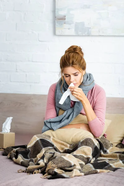 Femme attrayante et malade avec écharpe grise assise dans le lit et tenant une serviette — Photo de stock