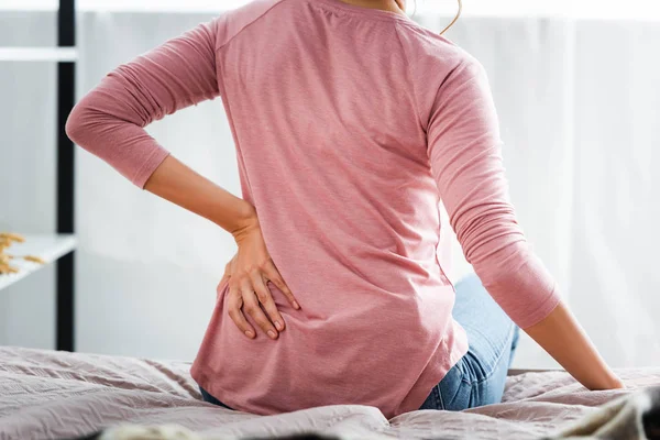 Vista posteriore della donna con dolore alla schiena seduta sul letto in appartamento — Foto stock