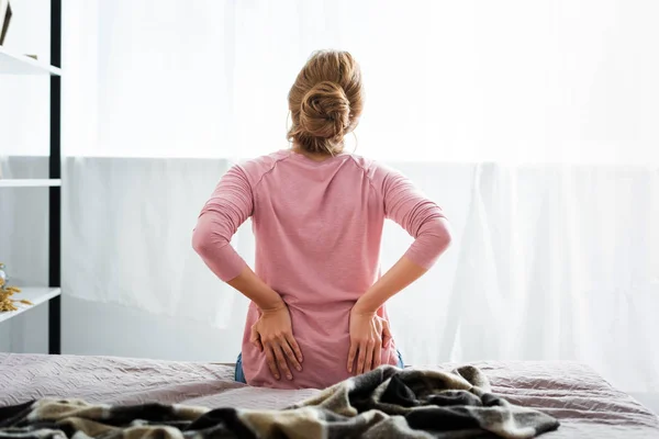 Вид ззаду жінки з болем у спині, сидячи на ліжку в квартирі — стокове фото
