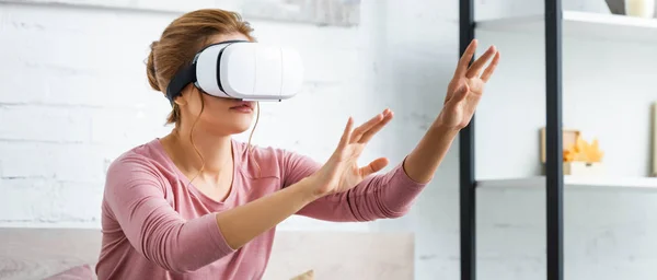 Панорамний знімок молодої дорослої жінки, що грає з гарнітурою віртуальної реальності в квартирі — стокове фото