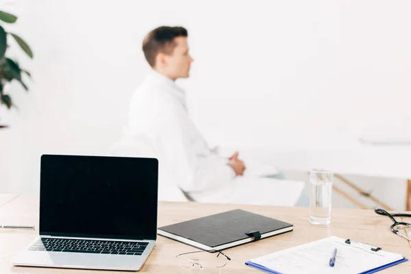 Selective focus of doctor in white coat and laptop with blank screen on foreground — Stock Photo