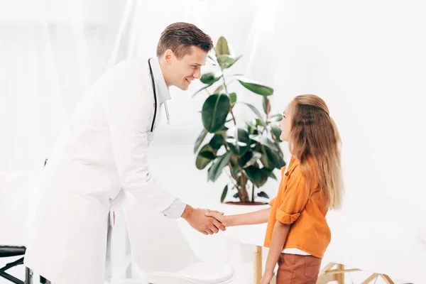 Pediatra con abrigo blanco y niño estrechándose la mano y mirándose - foto de stock