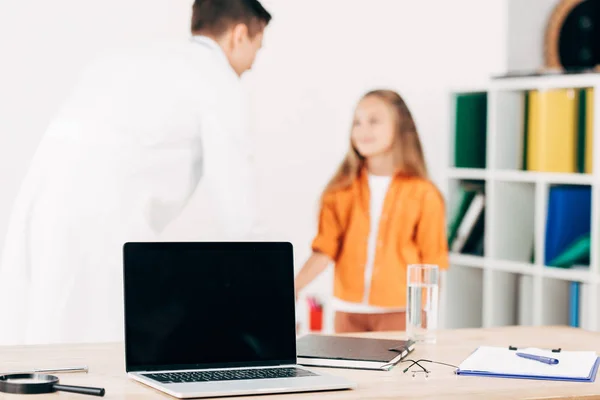 Foco seletivo do pediatra e paciente e laptop com tela em branco em primeiro plano — Fotografia de Stock