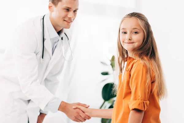 Pediatra con abrigo blanco y niño estrechándose la mano y mirándose - foto de stock