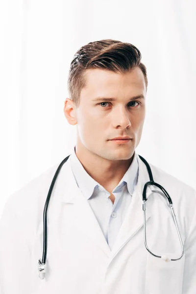 Médecin sérieux en manteau blanc avec stéthoscope regardant la caméra sur blanc — Photo de stock