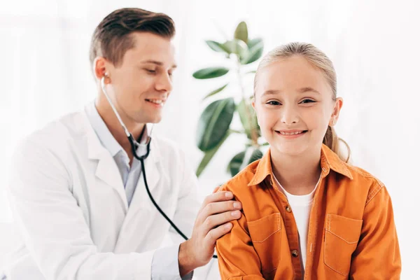 Улыбающийся педиатр в белом халате осматривает ребенка стетоскопом — стоковое фото