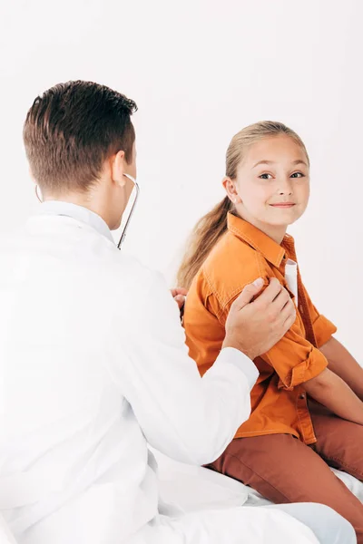 Педиатр в белом халате осматривает ребенка со стетоскопом изолированным на белом — стоковое фото