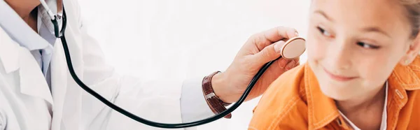 Panoramaaufnahme eines Kinderarztes im weißen Kittel, der ein Kind mit Stethoskop isoliert auf weißem Grund untersucht — Stockfoto