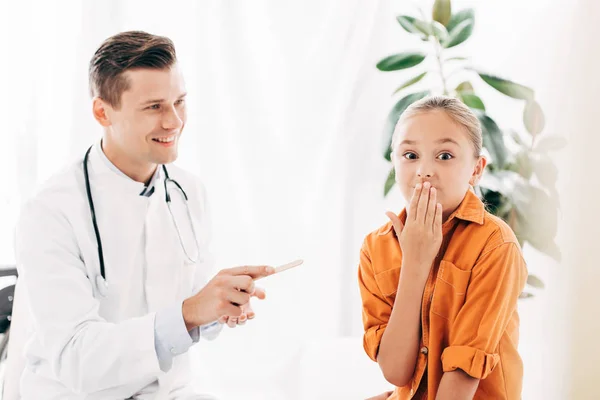 Criança cobrindo a boca com a mão e pediatra sorrindo segurando espátula médica na clínica — Fotografia de Stock