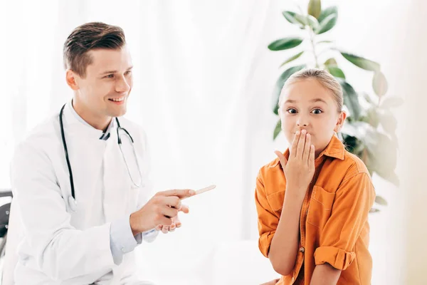 Kid covering mouth with hand and smiling pediatrist holding medical spatula in clinic — Stock Photo