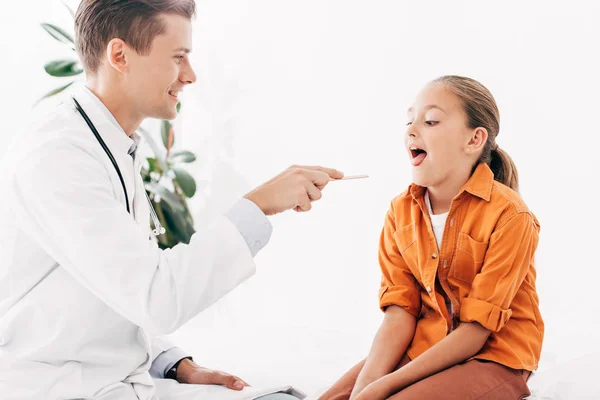 Pediatra de bata blanca examinando a un niño con espátula médica - foto de stock
