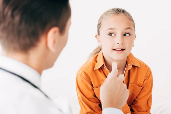 Enfoque selectivo del pediatra en bata blanca examinando al niño con espátula médica - foto de stock