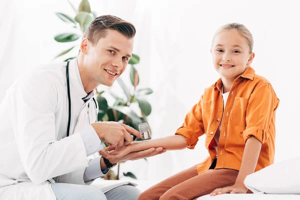 Pédiatre souriant en manteau blanc examinant l'enfant avec dermascope — Photo de stock