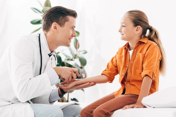 Улыбающийся педиатр в белом халате осматривает ребенка дермаскопом — стоковое фото