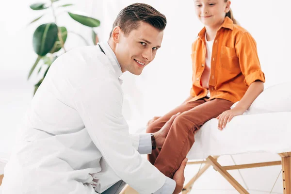 Улыбающийся педиатр в белом халате осматривает ребенка в клинике — стоковое фото