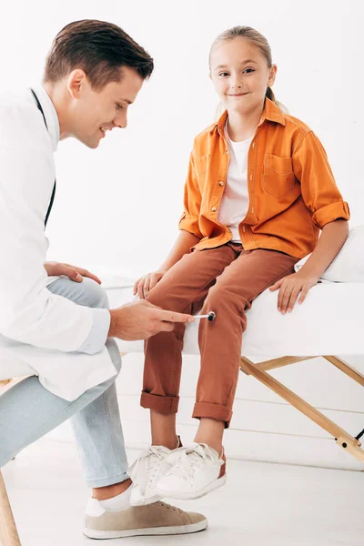 Pediatra sonriente en bata blanca examinando niño con martillo reflejo - foto de stock