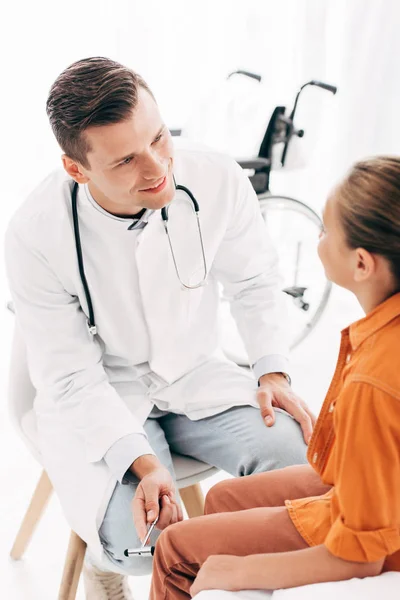 Pediatra sorridente em casaco branco examinando criança com martelo reflexo — Fotografia de Stock
